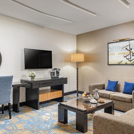 อัลคูฟราน ซาฟวะฮ์ โฮเต็ล มักกะฮ์ Hotel Mecca ภายนอก รูปภาพ The photo depicts a stylish hotel room or lounge area. It features a comfortable seating arrangement with two sofas and a coffee table in the center. There is a flat-screen television mounted on the wall, alongside a decorative piece of art. A small 