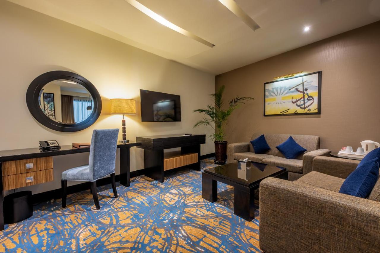 อัลคูฟราน ซาฟวะฮ์ โฮเต็ล มักกะฮ์ Hotel Mecca ภายนอก รูปภาพ The photo shows a hotel room or a lounge area. It features a seating arrangement with a light-colored sofa and a matching armchair. There is a coffee table in front, set on a patterned blue and orange carpet. A flat-screen television is mounted on th