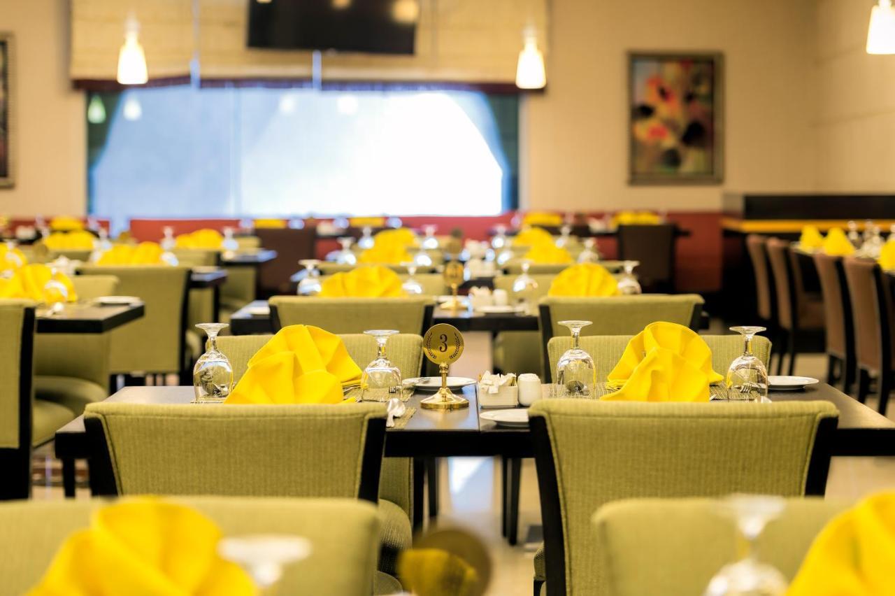 อัลคูฟราน ซาฟวะฮ์ โฮเต็ล มักกะฮ์ Hotel Mecca ภายนอก รูปภาพ The photo shows a restaurant interior with several dining tables arranged in a spacious setting. Each table is set with glasses, utensils, and folded yellow napkins. The chairs have a green upholstery, and there's ambient lighting with hanging fixtur