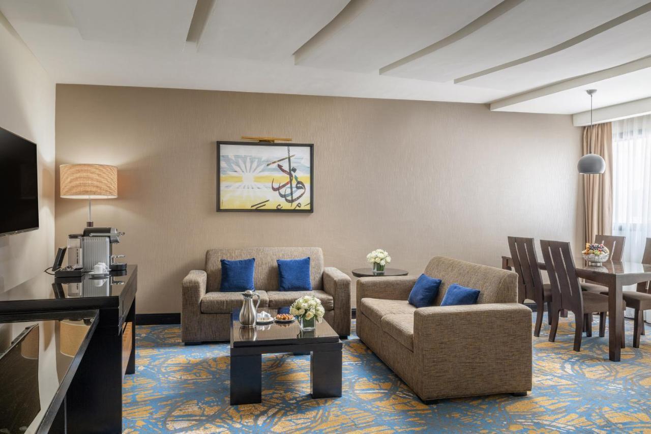 อัลคูฟราน ซาฟวะฮ์ โฮเต็ล มักกะฮ์ Hotel Mecca ภายนอก รูปภาพ The photo shows a well-decorated hotel suite or living area. It features a seating arrangement with two beige sofas, adorned with blue cushions. A small coffee table sits in front of the sofas, decorated with flowers. To the side, there's a dining ta