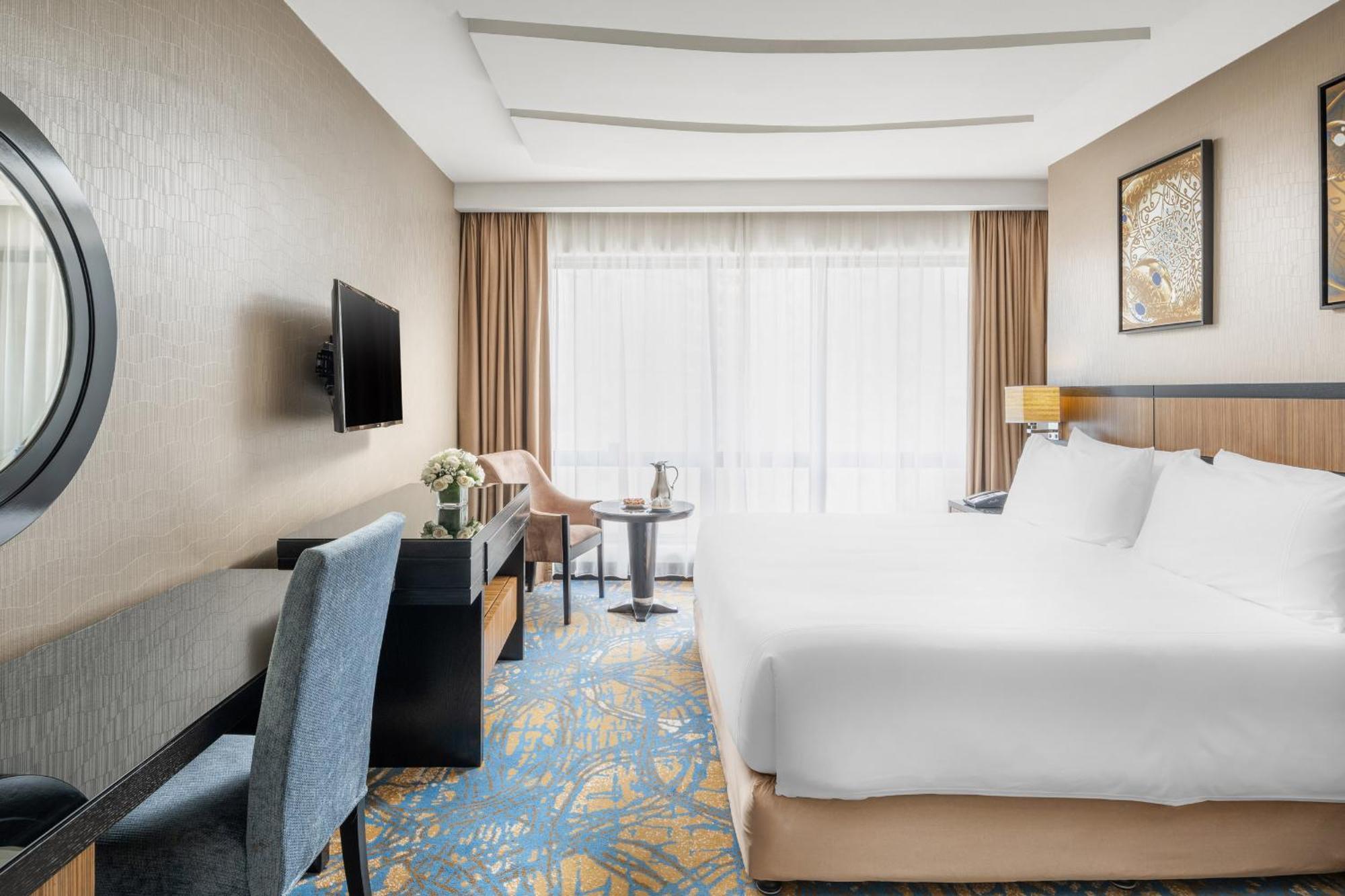 อัลคูฟราน ซาฟวะฮ์ โฮเต็ล มักกะฮ์ Hotel Mecca ภายนอก รูปภาพ The photo shows an elegantly designed hotel room. It features a large bed with white linens, a comfy chair, and a small table with two chairs. There is a television mounted on the wall and a decorative piece hanging nearby. The room has large windows