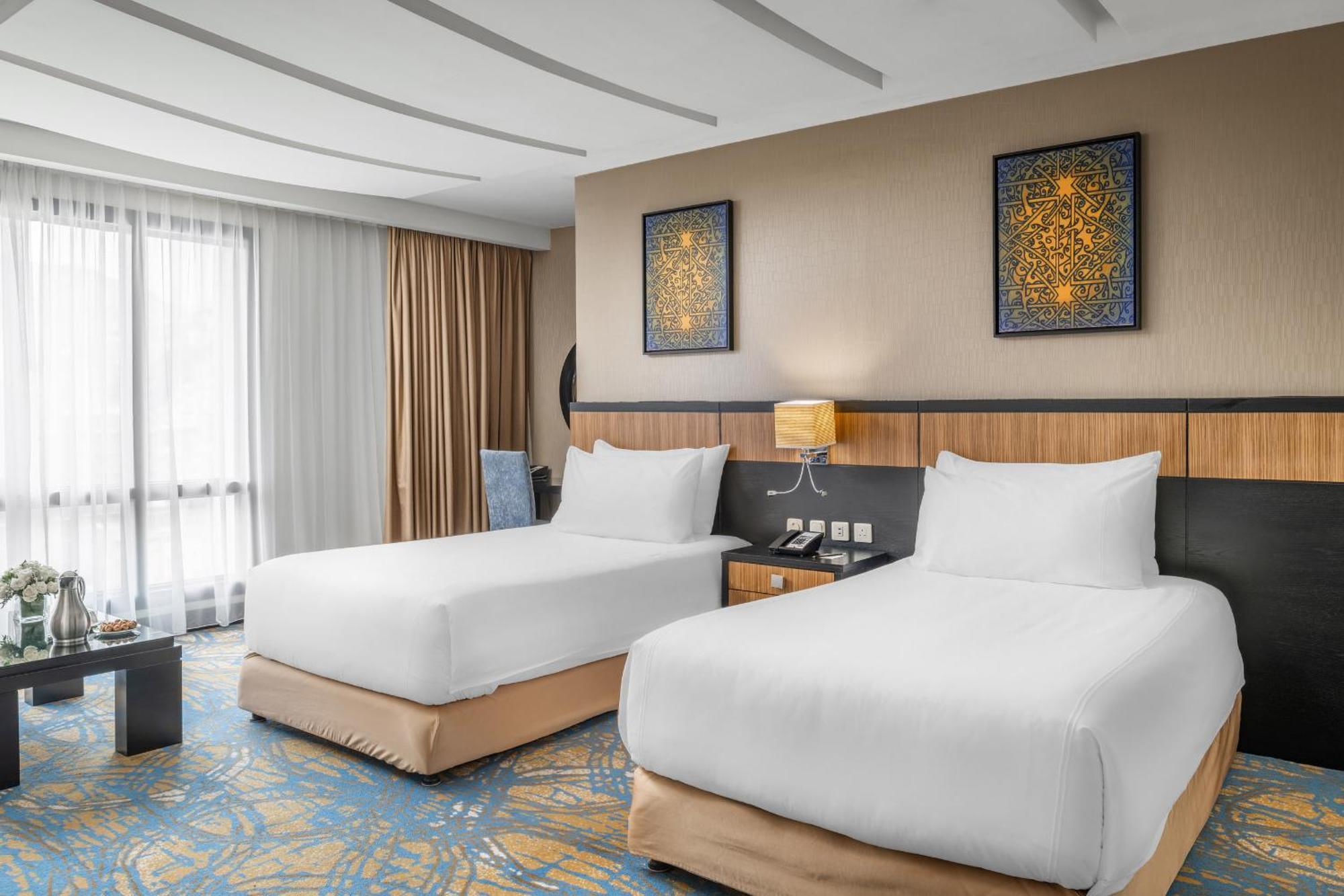 อัลคูฟราน ซาฟวะฮ์ โฮเต็ล มักกะฮ์ Hotel Mecca ภายนอก รูปภาพ The photo shows a modern hotel room featuring two white bedding twin beds with soft beige bases. There are decorative paintings on the wall above the beds, and a bedside table with a lamp is positioned between them. The room has large windows covered