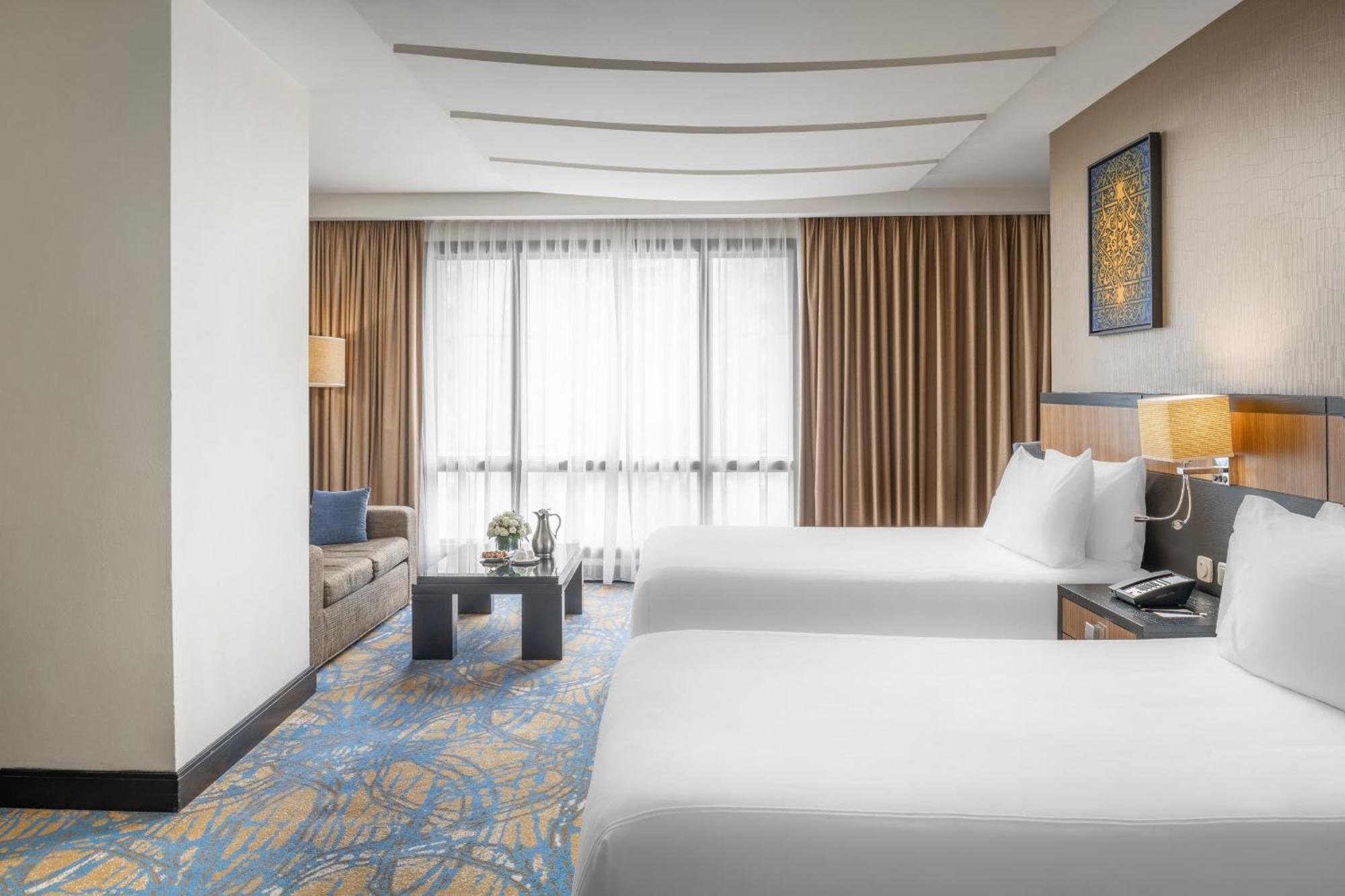 อัลคูฟราน ซาฟวะฮ์ โฮเต็ล มักกะฮ์ Hotel Mecca ภายนอก รูปภาพ The photo shows a hotel room with two comfortable beds, dressed in white linens. The decor features a warm color palette, with brown curtains and a stylish, contemporary design. There is a small sofa in the corner beside a coffee table, creating a co